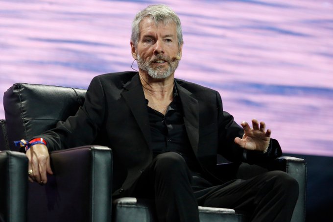 A gray-bearded man in a black suit sits in a chair with his hand outstretched, speaking on stage with a headset microphone.