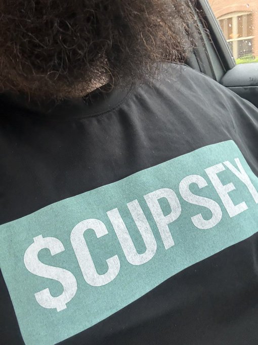 A person with curly hair wearing a black t-shirt featuring a large teal "$CUPSEY" logo across the chest, representing their support for the Cupsey token brand.