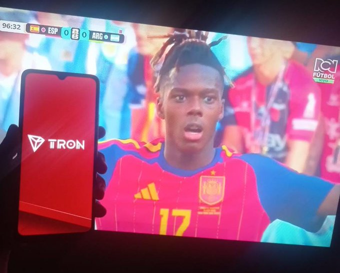 A hand holds a phone displaying the "TRON" logo in front of a TV broadcasting a Spain vs. Argentina match (ESP 0 - 0 ARG) featuring player #17 Nico Williams, visually pairing the TRON brand with the excitement of the global soccer tournament as suggested by the post's caption about uniting fans through fast, borderless payments.