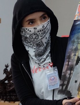 The image shows a person wearing a black hoodie and a black-and-white bandana covering their face, holding up what appears to be a piece of artwork. They have an ID badge visible on their chest, and the accompanying post connects them to their art available on @TransientLabs, suggesting the image showcases an artist displaying or promoting their creative work on the platform.