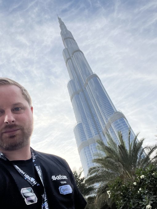 Selfie of a man wearing a 'SofTeam' jacket and a Hackathon lanyard in front of the Burj Khalifa in Dubai.