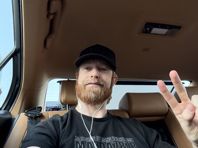 A selfie of a bearded man wearing a black cap and t-shirt with earbuds in, holding up a peace sign while sitting inside a vehicle with tan leather seats.