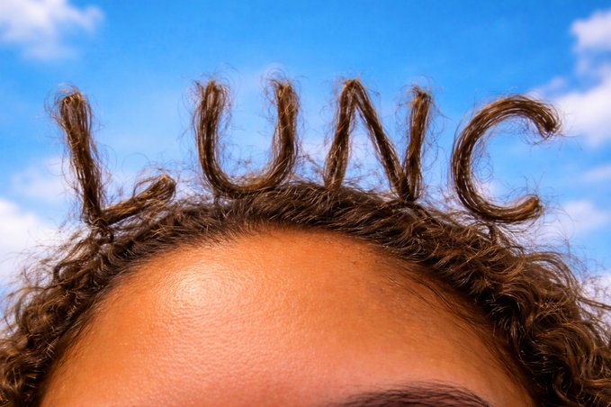 Close-up of a person's forehead and curly brown hair against a blue sky, with individual hair strands creatively arranged to spell 'LUNAC'.