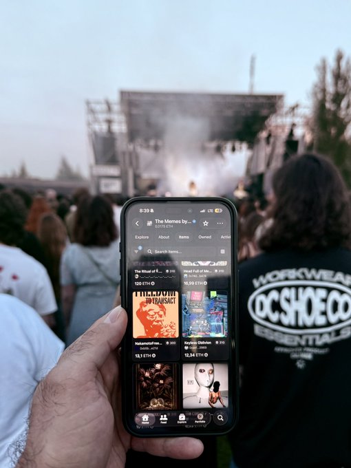 This is a snapshot of a person who appears to be standing at a music festival, taking a selfie while checking their phone. The phone screen displays the OpenSea app with various graphics of NFTs for sale.
