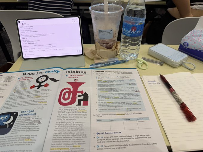 A study desk scene featuring an open grammar textbook (Oxford Grammar Bank 16), an iPad displaying code, a Starbucks iced coffee cup, a bottle of SeaMaster water, scissors, and various stationery items including highlighters and a small pouch.