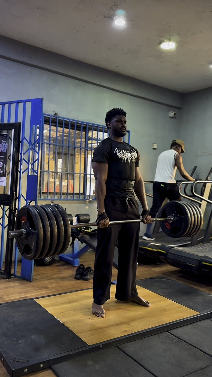 A man wearing a black t-shirt and weightlifting belt stands barefoot on a wooden lifting platform in a gym, holding a heavily loaded barbell. Another person walks on a treadmill in the background.