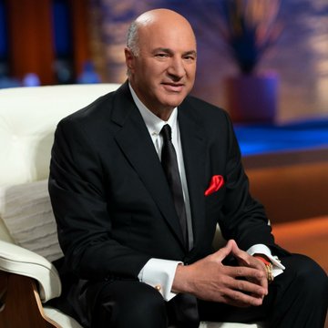 Billionaire Kevin O'Leary sits in a white chair wearing a dark suit with a red pocket square, smiling for a portrait.