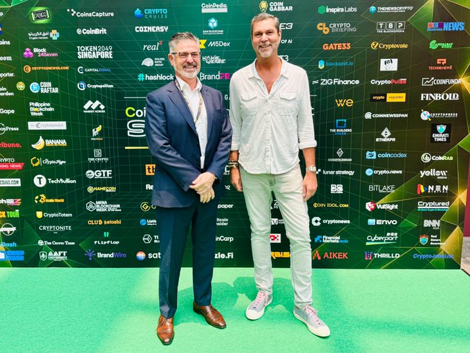 Two men posing together at a crypto/blockchain event, standing on a green carpet in front of a sponsor backdrop featuring logos such as CoinGabar, Coinbase, and CryptoBriefing.