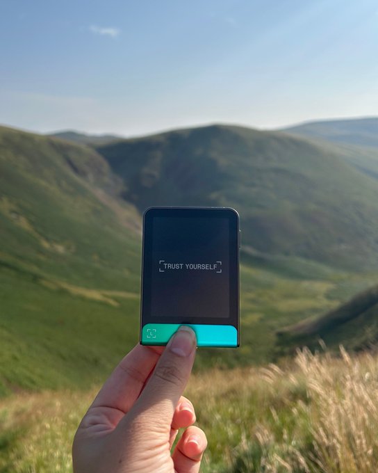 A hand holds up a small teal and black device displaying the words "TRUST YOURSELF" against a scenic mountain backdrop, perfectly capturing the "touch grass" spirit of enjoying a secure Sunday outdoors. The minimalist affirmation screen echoes the post's message of self-reliance and mindful presence in nature.