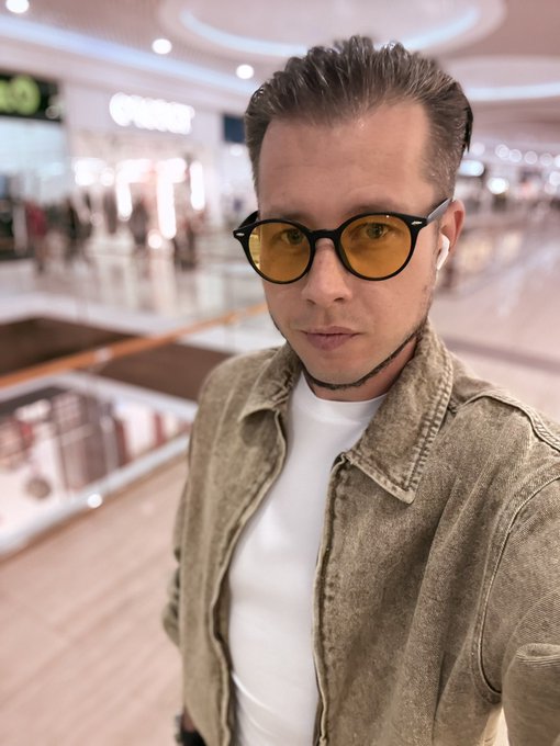 A selfie of a man wearing a denim jacket, white t-shirt, yellow-tinted sunglasses, and white wireless earbuds in a shopping mall.