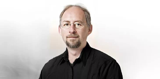 A portrait of Adam Back, the cypherpunk and Blockstream CEO, wearing glasses and a black shirt against a soft, hazy background. The image accompanies his quote on enduring Bitcoin market downturns by framing dips as wealth transfers to patient, long-term holders.
