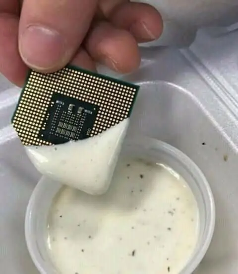 A hand dipping a computer processor chip (CPU) into a container of creamy liquid speckled with herbs or seeds.