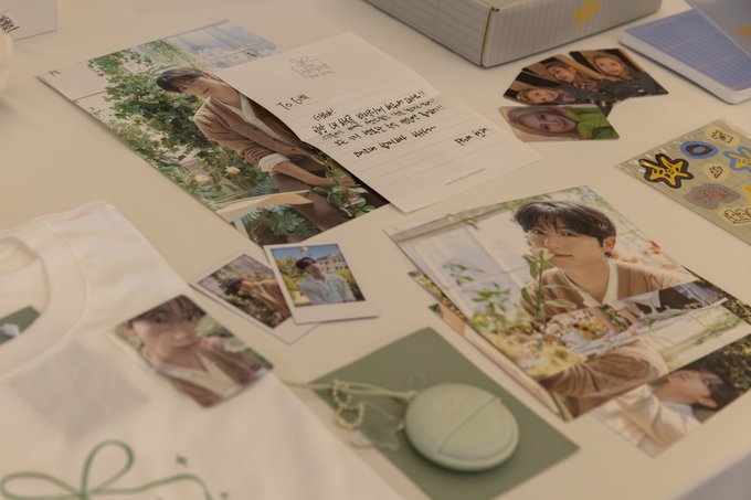 A flat lay of various merchandise including postcards, polaroids, a handwritten note, a tote bag, and a small round container.