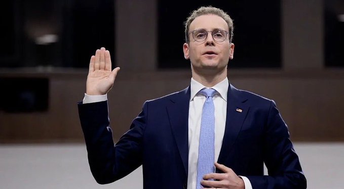 A man with glasses in a dark suit and light blue tie raises his right hand as if taking an oath or making a pledge, with an American flag pin on his lapel.