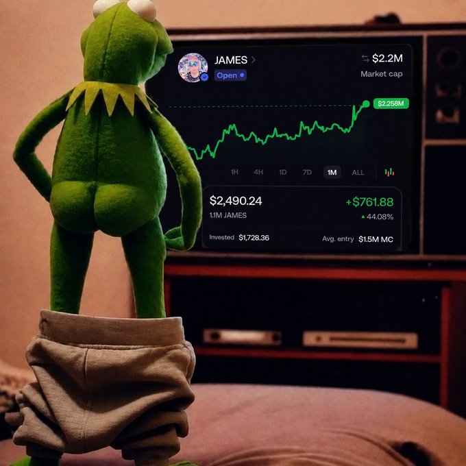 A Kermit the Frog plush toy sits on a bed facing a TV displaying a crypto trading chart for 'JAMES' with a market cap of $2.2M and a 1M position showing +44.08% profit.