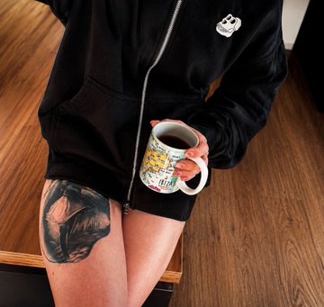 The image features a person sitting at a table with their legs crossed, holding a mug filled with coffee. The person has tattoos on their legs and arms, adding an artistic touch to their appearance. They are wearing a black hoodie over a white shirt, giving them a casual yet stylish look. In the background, there is a wooden dining table that adds warmth to the scene. The image also contains text elements related to the post, such as "Good Morning my dear’s It's beautiful to wake up early and realize that I'm a part of @MetaVixensNFT & @BoredApeYC Now drink some coffee from @Jungle_Brew_Co Breakfast is the most important meal of the day Enjoy your Humpday".