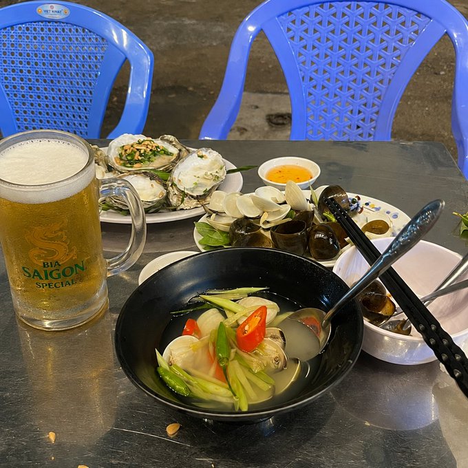 A table at an outdoor restaurant featuring a glass of 'BIA SAIGON SPECIAL' beer, a plate of oysters, and a bowl of clam soup with vegetables.