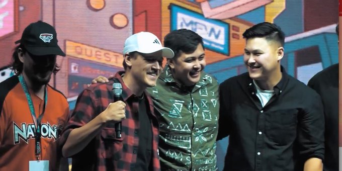 Photo of four men smiling together on stage, with one wearing a 'NATION' jersey and cap, another in a red plaid shirt holding a microphone, set against a colorful convention backdrop.