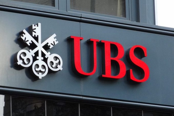 The image shows a UBS bank sign featuring the company's logo with three crossed keys alongside the bold red "UBS" lettering on a dark gray building facade. This signage directly relates to the post about UBS increasing its Bitcoin ETF holdings by 230%, highlighting the major financial institution's growing involvement in cryptocurrency investments.