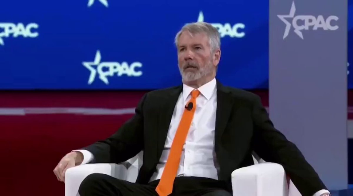 The image features a man dressed in a suit sitting comfortably in a chair, wearing an orange tie. He appears to be engaged in conversation or contemplation at some event. In front of him, there are blue stars displayed, which might signify the presence of an audience or prominence during his speech. The text at the bottom of the image is not legible, and it seems unrelated to the suit-wearing man in the chair with a tie.
