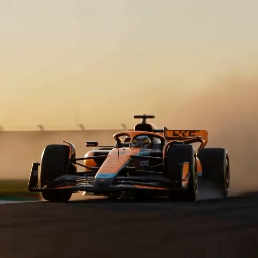 A McLaren Formula 1 race car speeds along a track at sunset, its sleek orange and black livery contrasting against the hazy, golden-lit backdrop. The image's high-speed racing theme mirrors the post's competitive trading competition, where six teams battle it out on a metaphorical "track" to be the fastest contributor in the race.
