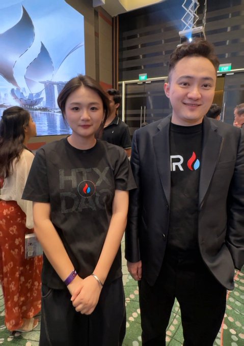 Two people pose together at an event, wearing matching 'HOX DAO' and 'R' branded black shirts, standing in front of a screen showing a lotus flower design.