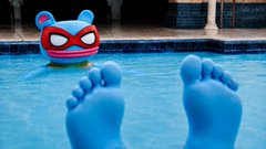 A surreal image showing a person's blue-tinted feet sticking out of a swimming pool, with a cartoon masked emoji floating in the water in the background.