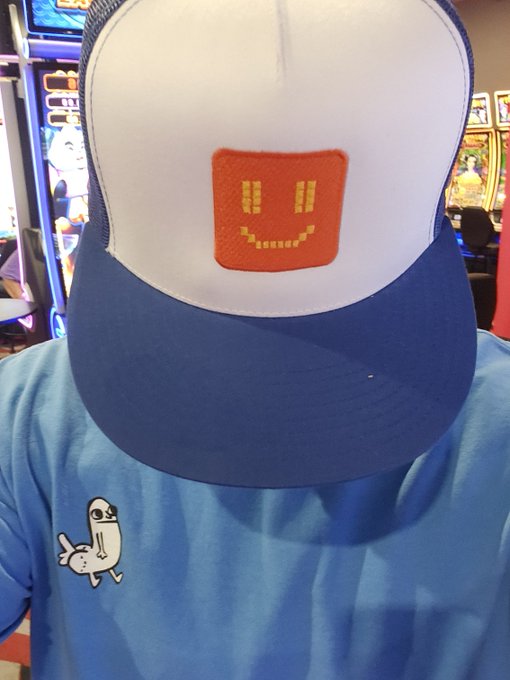 A close-up of a blue and white trucker cap featuring an orange smiley-face patch, with the wearer also sporting a blue shirt adorned with a small cartoon character, captured in what appears to be a casino setting. The "$BNKR mfer 4 life" caption suggests the poster identifies with the Bankr brand/meme aesthetic, tying the playful smiley imagery to their loyalty as a $BNKR token holder.