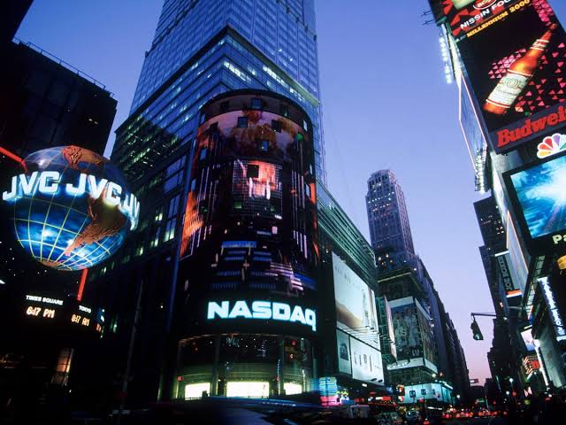 Times Square's towering NASDAQ building glows at dusk, prominently displaying the "JVC JVC" logo and "NASDAQ" signage, symbolizing the traditional financial markets now extending into round-the-clock trading hours.