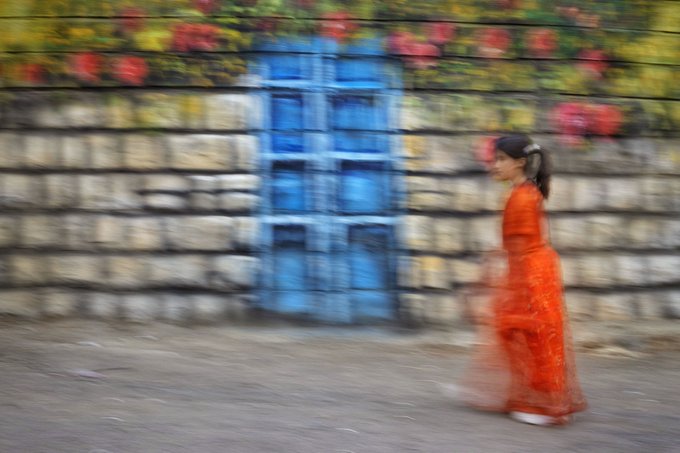 A motion-blurred photograph of a woman in a flowing orange dress walking past a stone wall with a blue door and red flowers above.