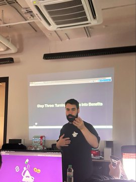 A man with a beard, wearing a black t-shirt, stands in a room with a ceiling-mounted projector screen displaying text about 'Xbox Series: TurnOn Controller Haptic Benefits', with a purple mascot visible on a monitor to the left.