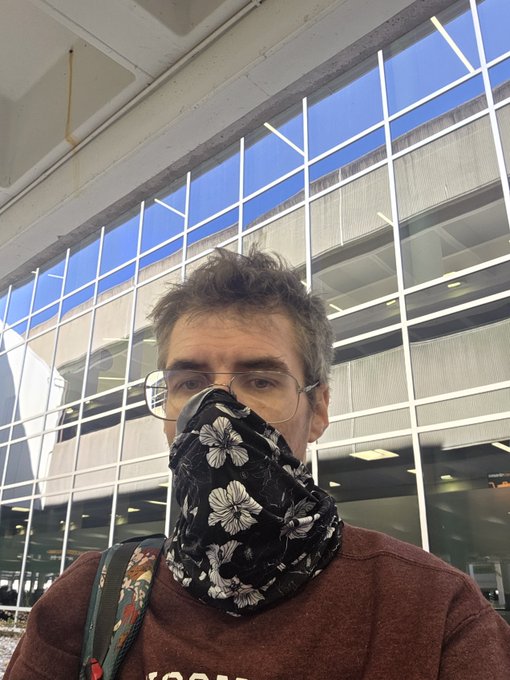 A selfie of a man wearing aviator glasses and a black floral bandana covering his face, standing in front of a modern glass building with a parking garage visible.