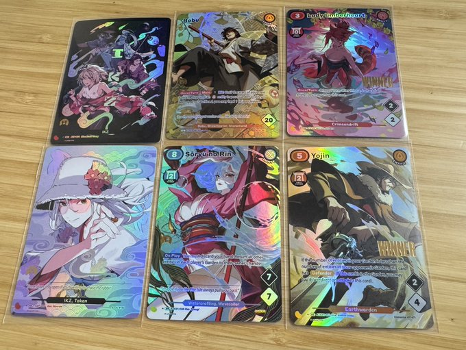 A collection of six holographic trading cards displayed on a wooden surface, featuring anime-style characters including Robin, LadyTimelesshcard, Yojin, SoryuuorRin, and characters labeled 'Earthwdern', with numerical stats visible on the cards.