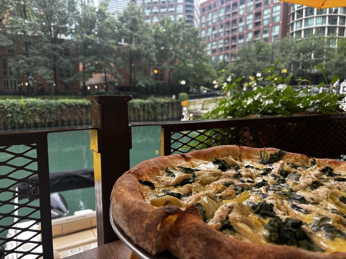 A spinach and artichoke pizza on a plate at an outdoor restaurant patio overlooking a green river and city buildings, likely along a Chicago waterway.