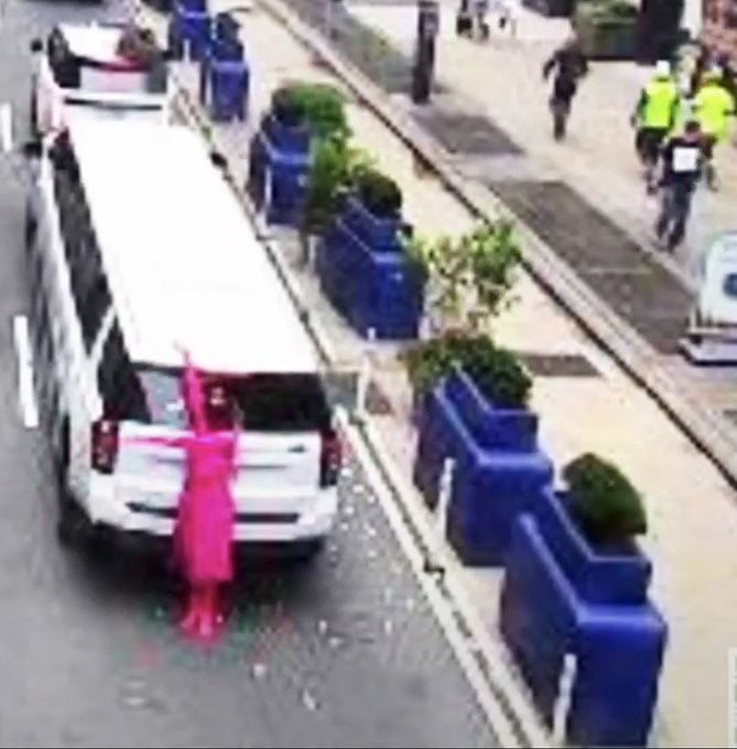 A low-resolution image showing a person in a bright pink dress standing at the open trunk of a white limousine parked along a blue-barriered street, with pedestrians in the background.