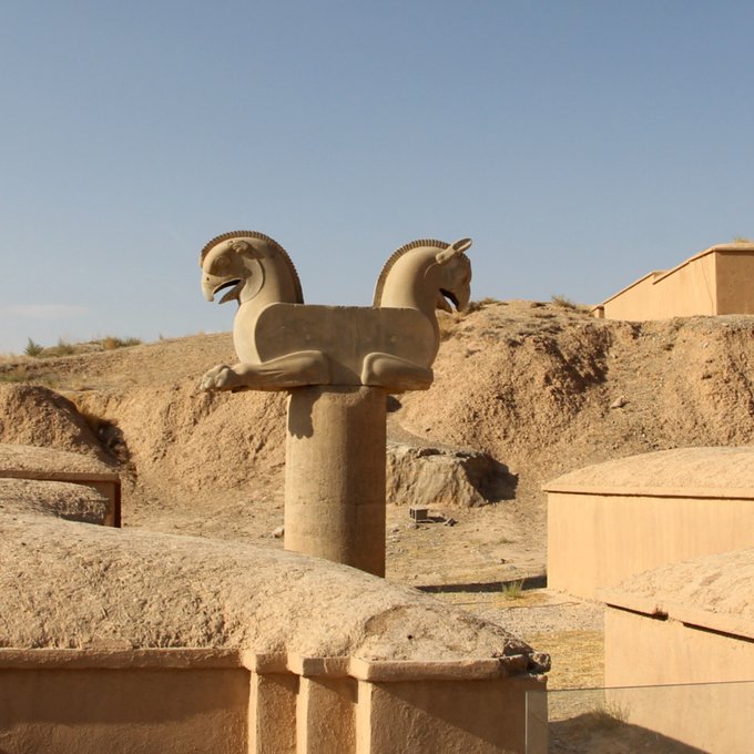 A stone statue of a double-headed creature (possibly a griffin or similar mythological beast) standing on a pillar in an arid, desert-like archaeological site.
