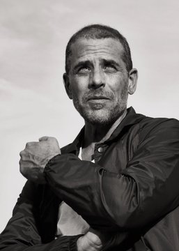 A black-and-white portrait photograph of Hunter Biden looking off to the side with a serious expression, wearing a dark jacket with one hand raised near his chest; the image accompanies a news post stating that he denied earning a "single dollar" from his $LAPTOP memecoin while the top 10 wallets reportedly profited over $2 million from it.