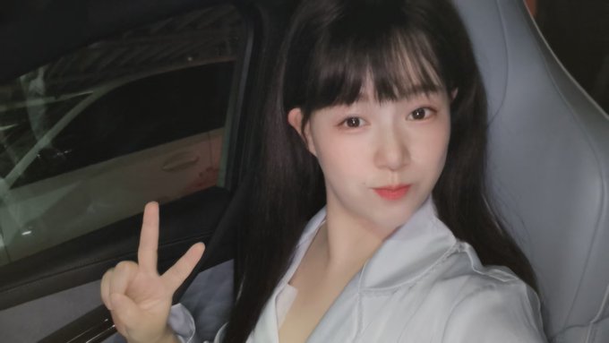 A young woman with long dark hair poses inside a car, making a peace sign with her hand while wearing a light-colored jacket.