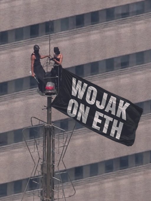 Two people stand atop a tall pole structure, hoisting a large black flag that reads "WOJAK ON ETH," symbolically planting their banner high above a city building to emphasize the project's Ethereum-based presence in line with the post's "to infinity & beyond" message for $WOJAK.