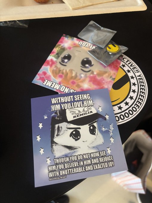 Religious-themed stickers and cards on a dark surface, including one reading "WITHOUT SEEING HIM YOU LOVE HIM" featuring Remilia and another with "GOD BLESS" and a smiling bread character.