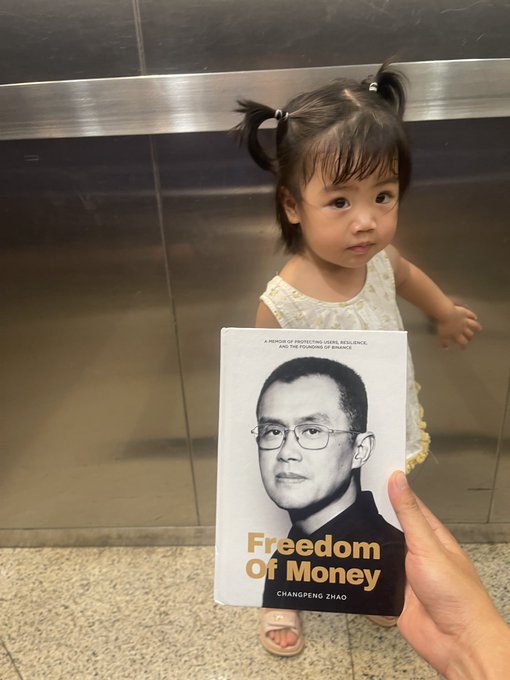 A toddler with pigtails holds up the book "Freedom of Money" by Changpeng Zhao in an elevator, accompanying a post about the author's hope to introduce her nearly 2-year-old daughter to the concepts of money, investing, and freedom through this book.