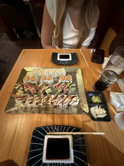 A large platter of assorted sushi rolls sits on a wooden table in front of a woman, accompanied by soy sauce dishes, wasabi, ginger, and chopsticks.