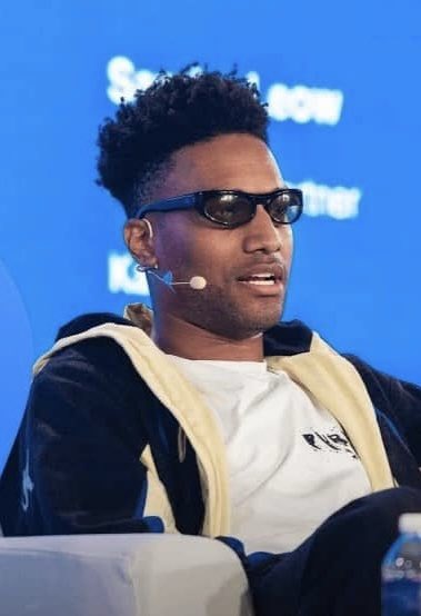 The image shows a man wearing dark sunglasses and a beige and black hoodie, speaking at what appears to be a conference or panel event with a headset microphone. Behind him, partial text on a blue backdrop reads "Show" and "nter," suggesting a "Show Center" or similar venue branding, fitting the context of the post about ANSEM making bullish remarks that "you are not getting lower prices."