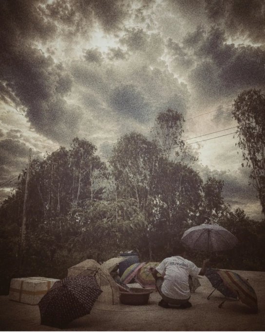An artistic photo showing a person crouched on the ground sorting items in baskets under several umbrellas, with dramatic cloudy skies and tall trees in the background.