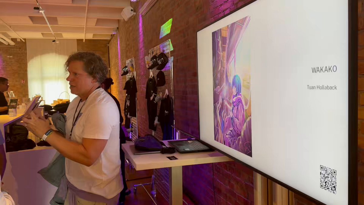 The image shows a woman standing next to a large screen displaying an art exhibit at a convention or event. The artwork features a painting of a person with purple hair. There are several people present, including the woman who appears to be reading something on her phone. A laptop is also visible in the scene, possibly used by someone to view more information about the exhibit or to take notes during the event.