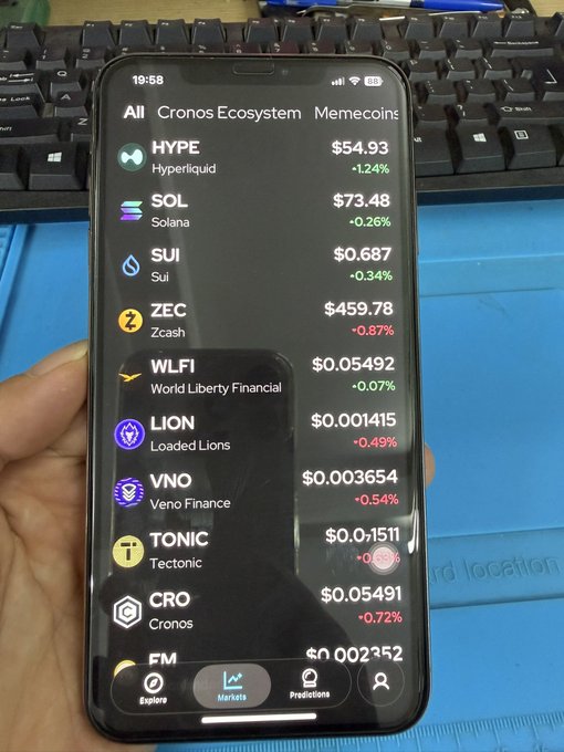 A smartphone displaying a crypto trading app showing prices for HYPE ($54.93), SOL ($73.48), SUI ($0.687), ZEC ($459.78), WLFI ($0.05492), LION ($0.001415), VNO, TONIC, CRO, and FM, held in front of a keyboard.