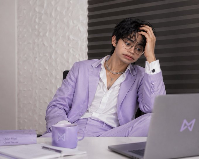 A young man with dark hair and round glasses wearing a light purple suit over a white shirt, sitting at a desk with a laptop and mug, with books labeled 'Quiet Mind Clear Future' nearby.