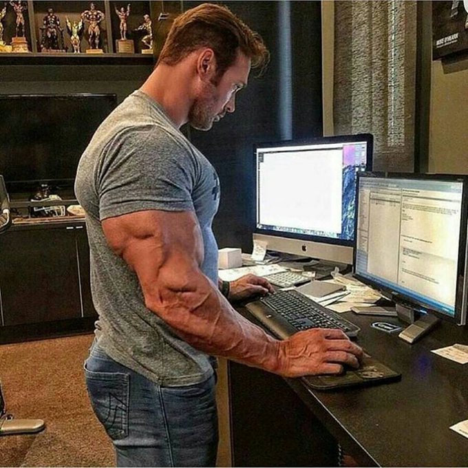 A muscular man in a gray t-shirt and jeans stands at a standing desk working on dual monitors, with action figures visible on shelves in the background.