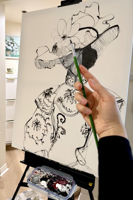 A photograph of an artist working on a black and white fashion illustration of a woman wearing an elaborate hat and floral dress, holding a green brush with a palette visible below.
