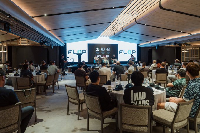 A view from the back of an audience seated at round tables in a conference room, facing a stage where panelists are seated in front of screens displaying 'FLOP'.